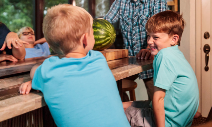 young boys smiling and sitting at table with adults in background with watermelon ready to be cut