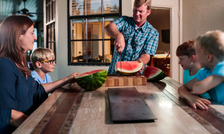 watermelon being cut for family by man with three boys and one woman at table