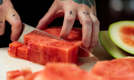 watermelon cubes being cut. hand and knife shown in process of cutting, cut rind on side