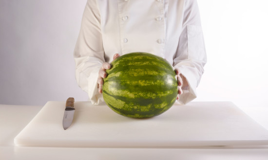 mini watermelon on cutting board with knife on side. person in chef coat in background holding watermelon on board