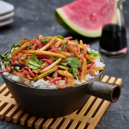 rind stir fry on top of white rice in small cast iron pan on top of bamboo trivet. serving plates and wedge of watermelon and cruet of soy sauce in background