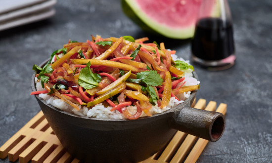 rind stir fry on top of white rice in small cast iron pan on top of bamboo trivet. serving plates and wedge of watermelon and cruet of soy sauce in background