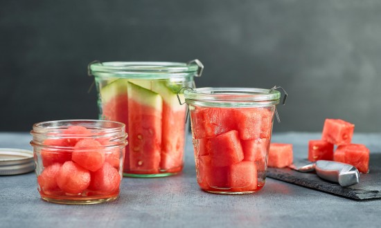 jars of watermelon cuts, balls, sticks, chunks, with knife and chunks in background. slate cutting board on side