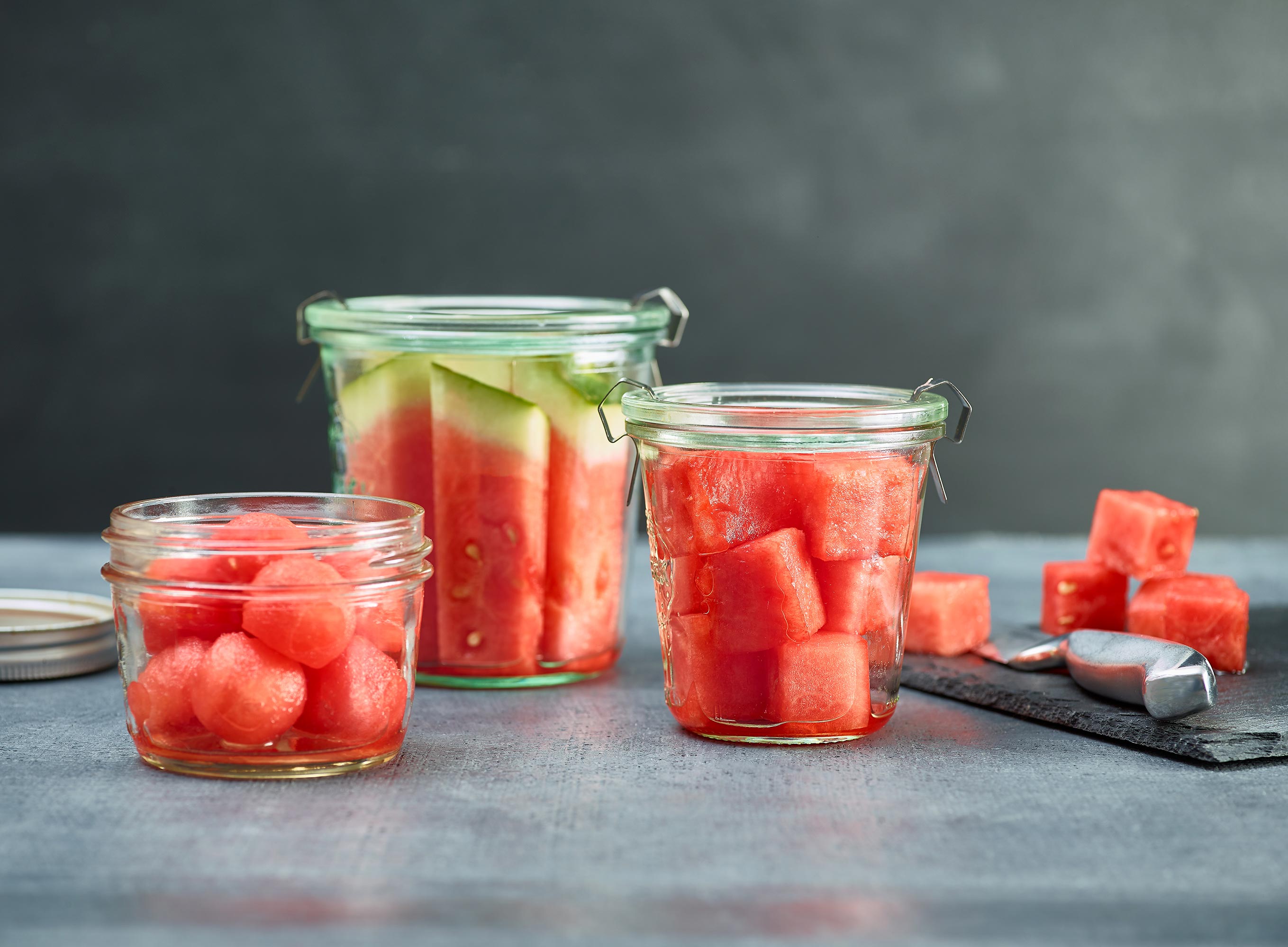 jars of watermelon cuts, balls, sticks, chunks, with knife and chunks in background. slate cutting board on side
