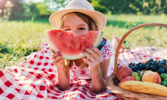 young girl on picnic red and white checkered tablecloth on ground holding watermelon slice. basket of fruit on side