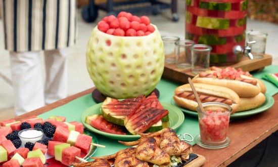 Celebrate Dad with Grill and Golf, Father's Day, menu items on wooden board on top of two round grills with cook in background holding plate of watermelon slices