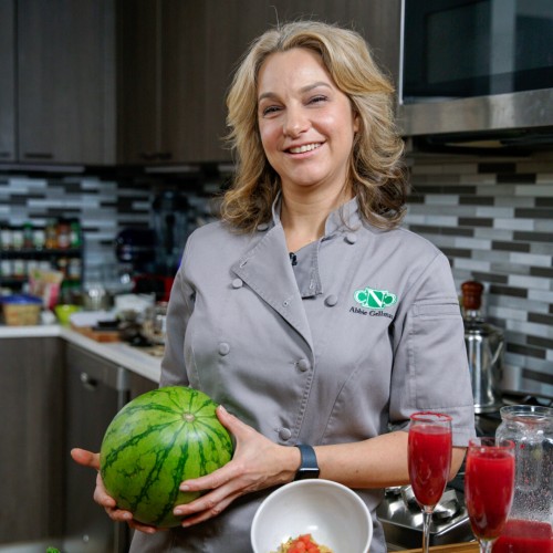 Abbie with watermelon in kitchen, holding mini watermelon. Oval platter with watermelon cuts, balls, strawberries, chocolate and two watermelon cocktails on side.