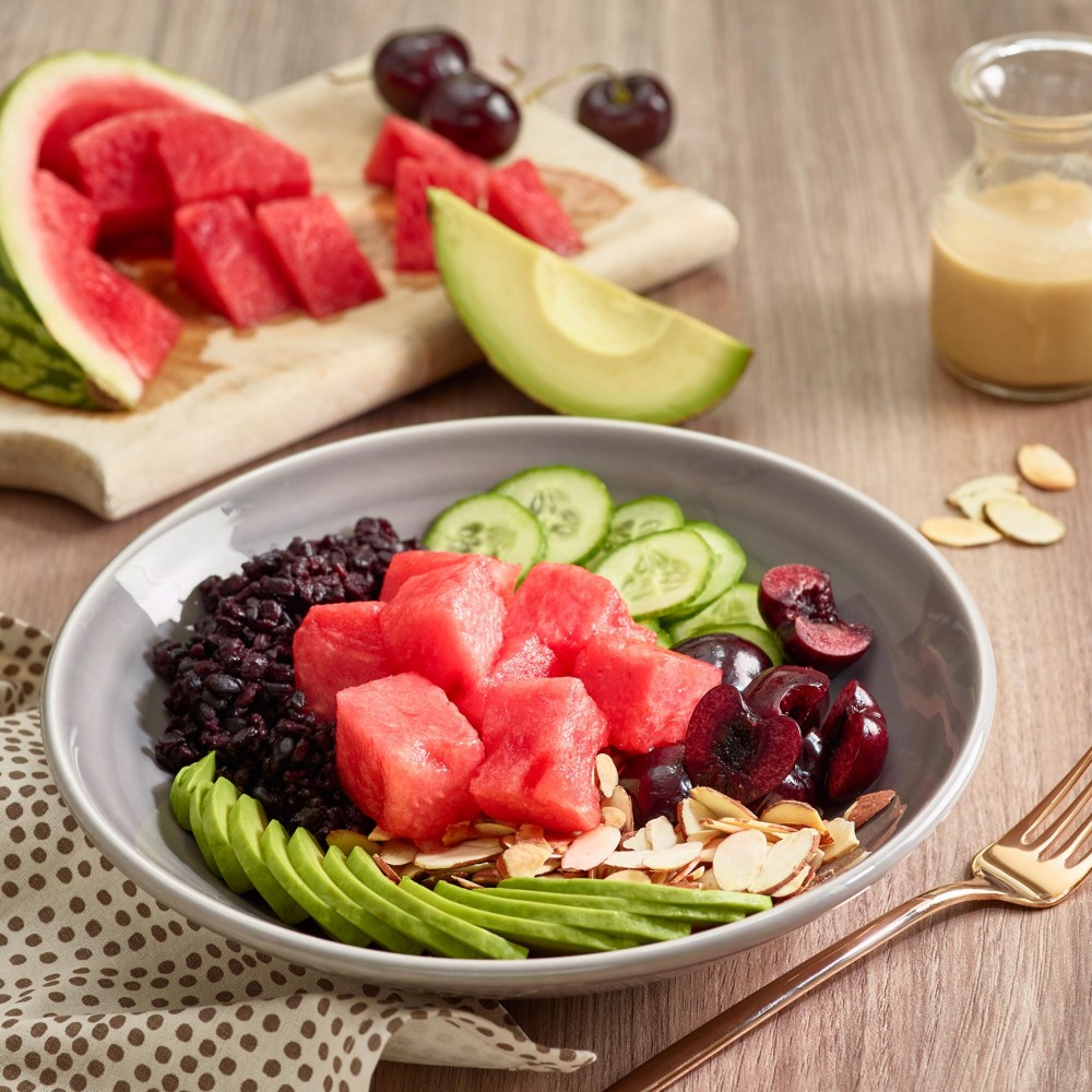 Buddha Bowl meal in gray ceramic bowl with watermelon chunks, cherries and avocado in background. Dressing on side in small cruet