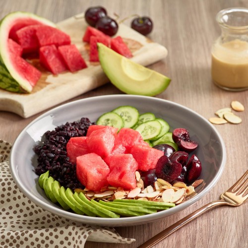 vegetarian meal in gray ceramic bowl with watermelon chunks, cherries and avocado in background. Dressing on side in small cruet
