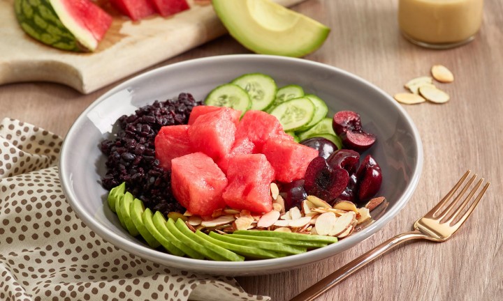 Buddha Bowl meal in gray ceramic bowl with watermelon chunks, cherries and avocado in background. Dressing on side in small cruet