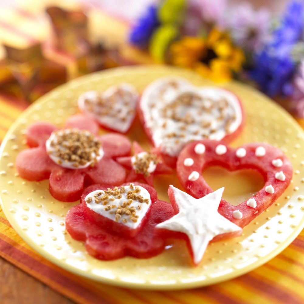 Frosted Watermelon Cutouts set on yellow plate with cutout cutters in background, also flowers in background