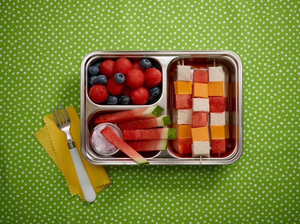 Happy Lunchbox - tin divided serving lunchbox with kebabs, watermelon balls and blueberries and sticks with yogurt dip. Fork on side on yellow napkin. Set on green with white polka dot background