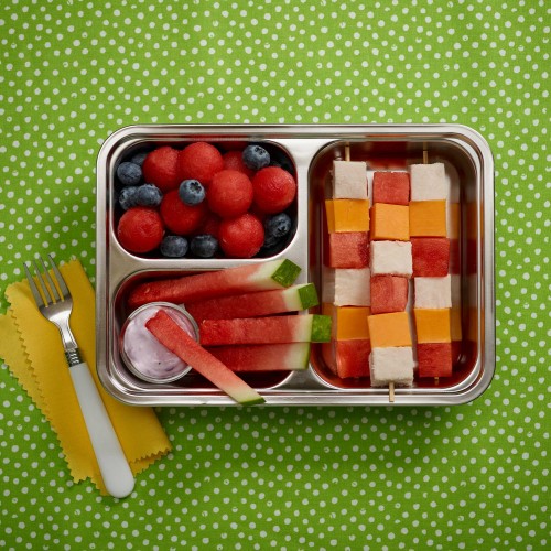 Happy Lunchbox - tin divided serving lunchbox with kebabs, watermelon balls and blueberries and sticks with yogurt dip. Fork on side on yellow napkin. Set on green with white polka dot background