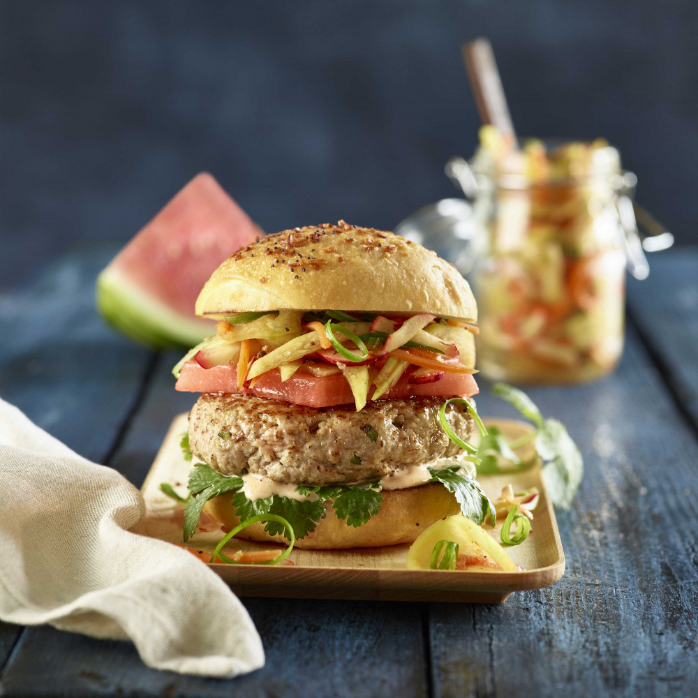 Kimchi Burger on rectangular bamboo plate with watermelon wedge and kimchi in jar with spoon in background, set on wooden table