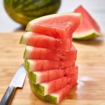 Stack of triangular wedges of red watermelon with larger wedge and mini melon in background. Knife next to cuts on bamboo cutting board