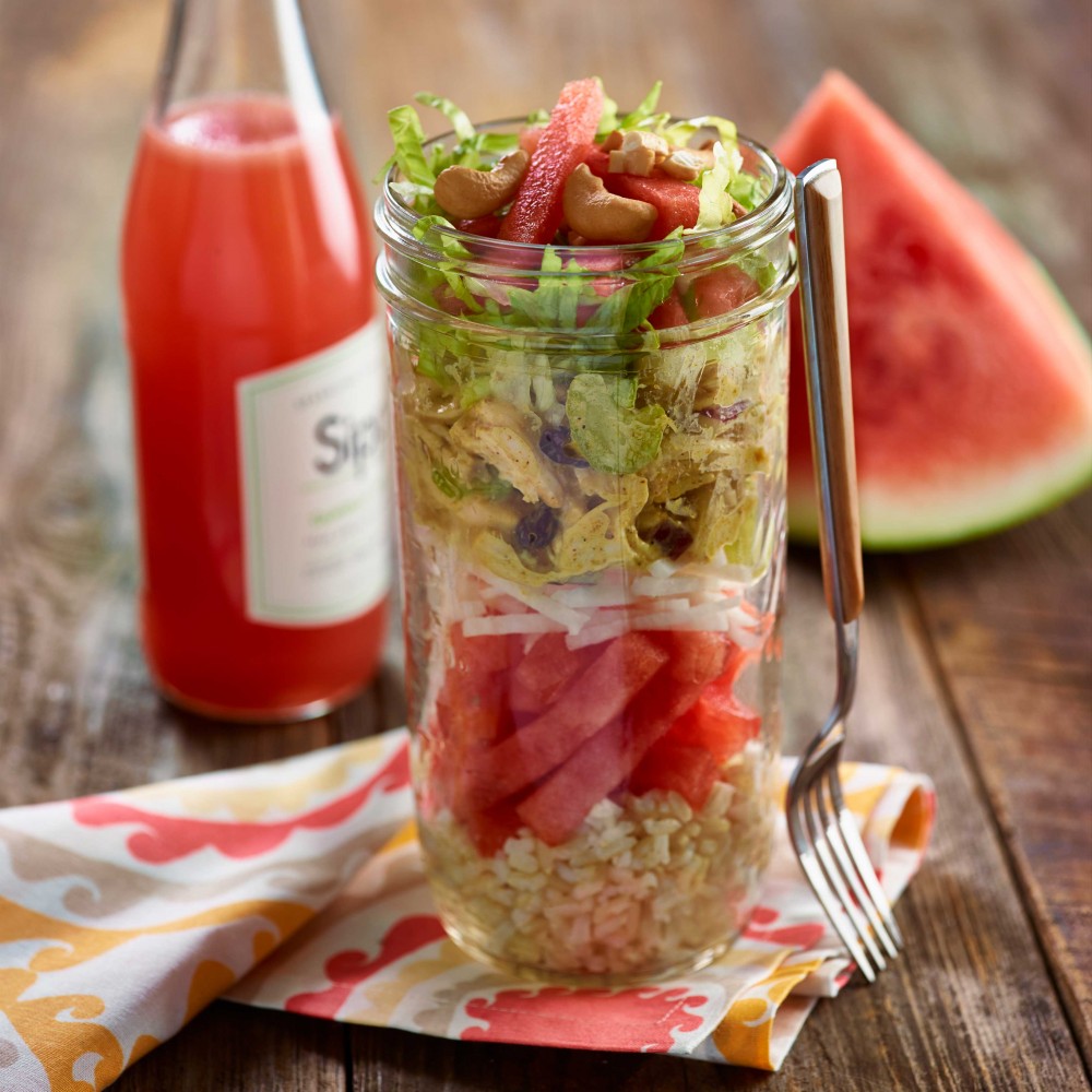 Stacked Jicama Chicken Salad stacked in tall clear jar on colorful napkin on wooden table. Watermelon drink and watermelon wedge in background