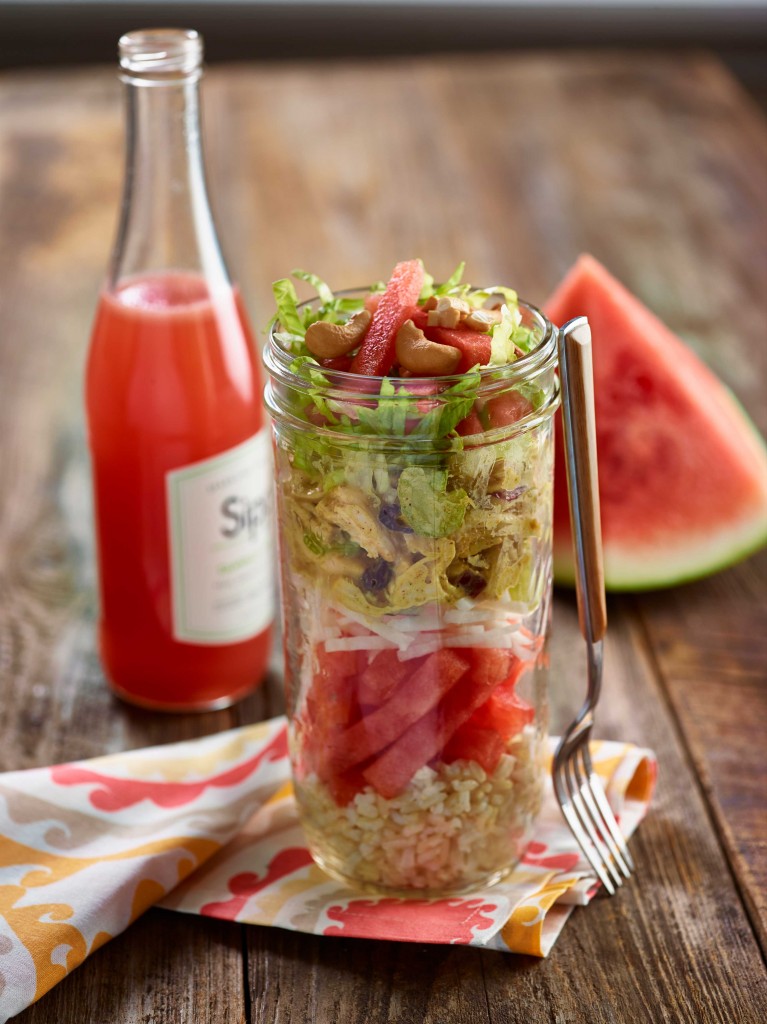 Stacked Jicama Chicken Salad stacked in tall clear jar on colorful napkin on wooden table. Watermelon drink and watermelon wedge in background