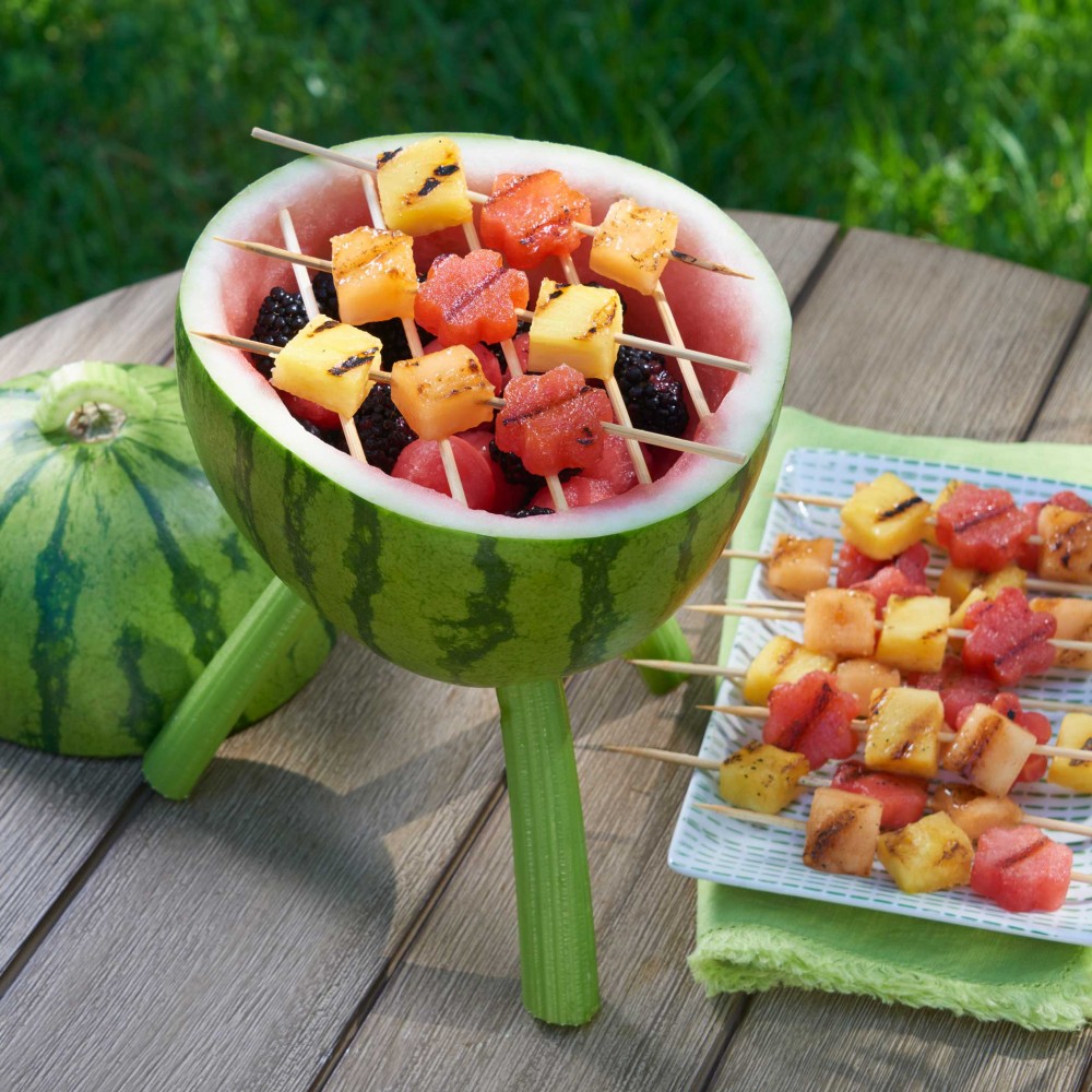 Watermelon Grill - carving with skewers of fruit on top of "grill" and some on appetizer plate set on green cloth napkin. Setting on small round wooden outdoor table. Grass in background