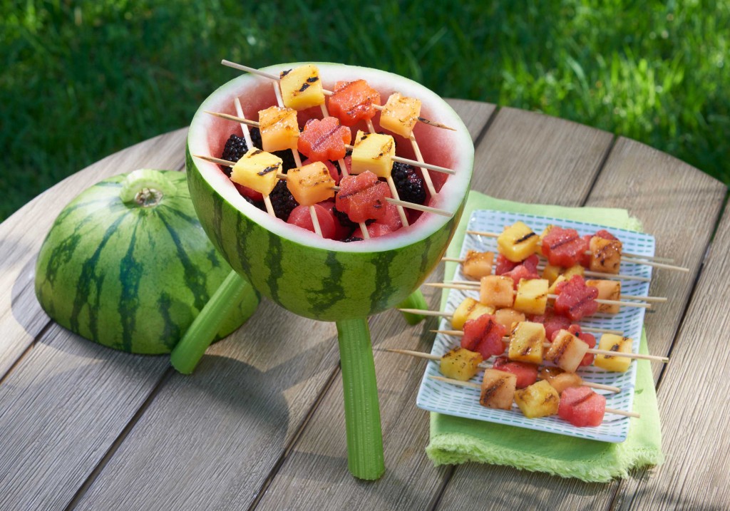 Watermelon Grill - carving with skewers of fruit on top of "grill" and some on appetizer plate set on green cloth napkin. Setting on small round wooden outdoor table. Grass in background