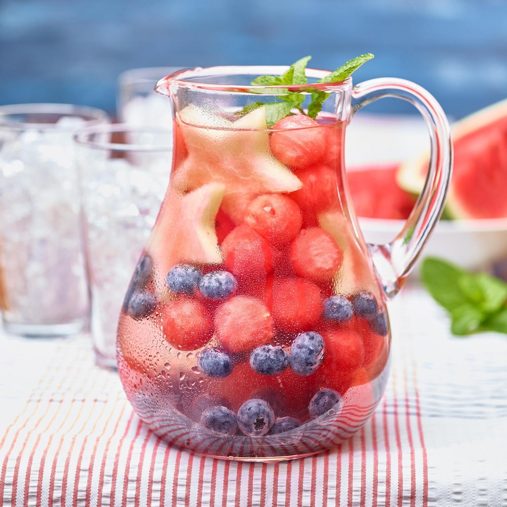 Watermelon Infused Water in pitcher filled with fruit (watermelon balls, blueberries, mint garnish. Clear glasses filled with crushed ice in background, also watermelon wedge in background. Set on red and white thin striped tablecloth