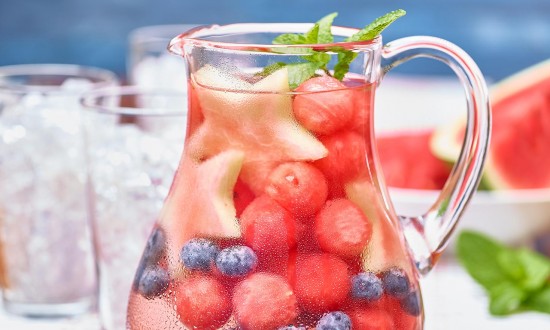 Watermelon Infused Water in pitcher filled with fruit (watermelon balls, blueberries, mint garnish. Clear glasses filled with crushed ice in background, also watermelon wedge in background. Set on red and white thin striped tablecloth