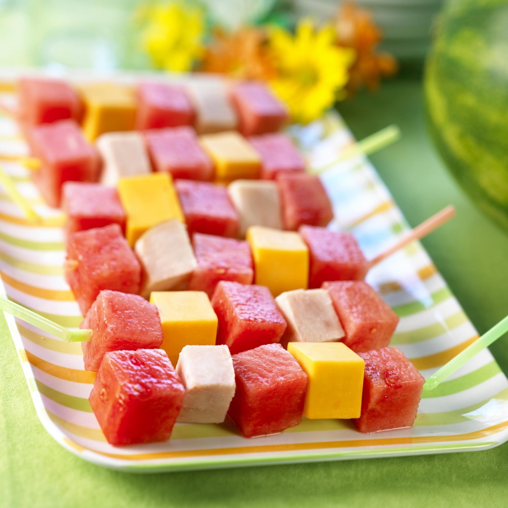 Watermelon Kebobs set on citrus colored striped oval serving tray with flowers and whole watermelon in background