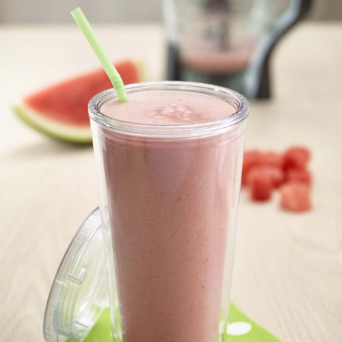 Watermelon Oatmeal Smoothie jin tall plastic glass with straw and lid on side. Blender, wedge of watermelon and chunks in background. Set on small green and large white polkadot paper napkin on wooden table