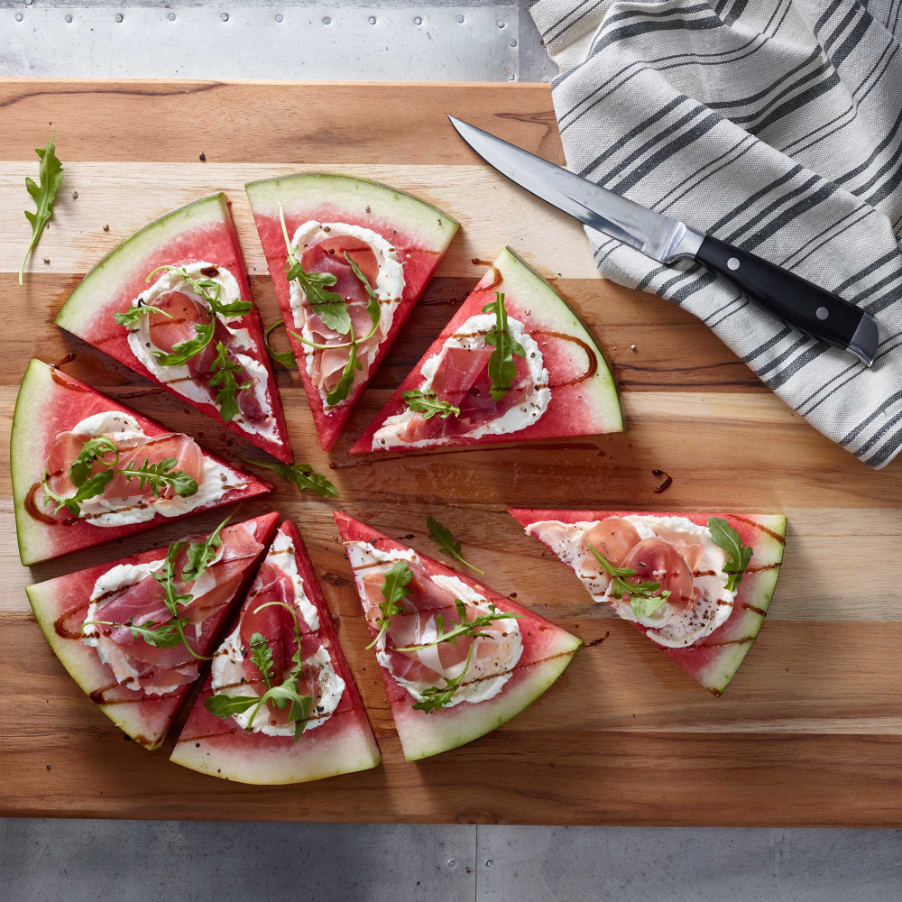 Watermelon pizza, savor shown on wooden cutting board, garnished with arugula, Knife on side of pizza with gray and white striped cloth