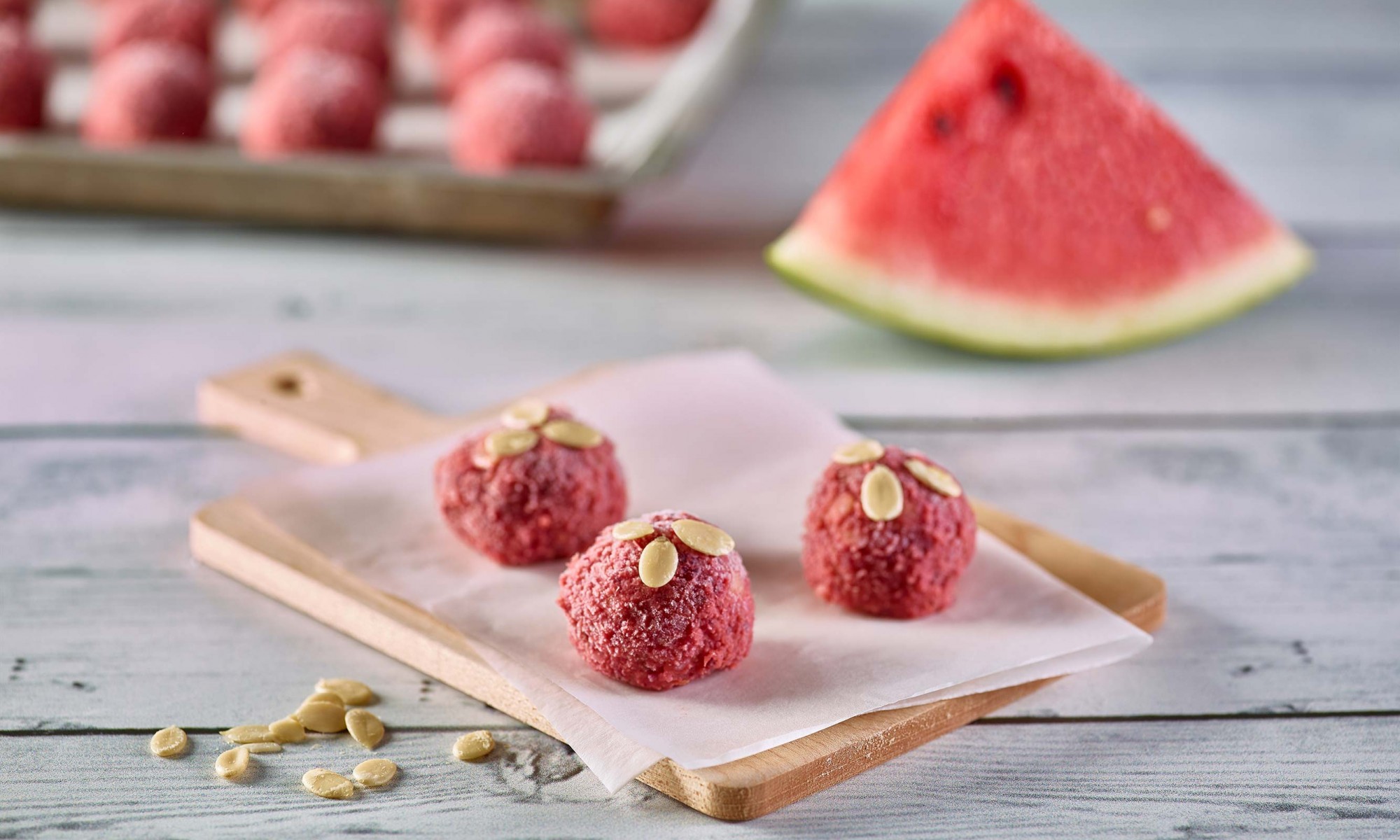 Watermelon Protein Bites set on parchment paper on small cutting board, garnished with watermelon seeds. Tray of more protein bites and wedge of watermelon in background
