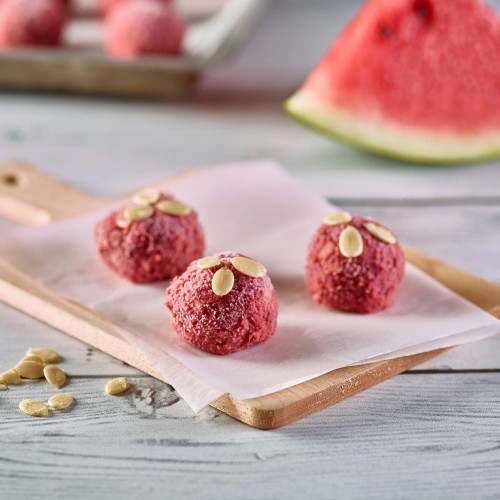Watermelon Protein Bites set on parchment paper on small cutting board, garnished with watermelon seeds. Tray of more protein bites and wedge of watermelon in background