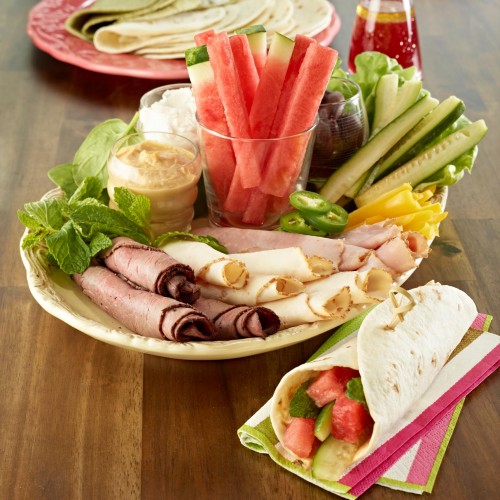Watermelon Sandwich Wraps set on serving platter with ingredients. Another plate in background with wraps. Whole watermelon and serving plates in background. One ready-to-eat wrap in foreground