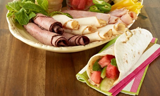 Watermelon Sandwich Wraps set on serving platter with ingredients. Another plate in background with wraps. Whole watermelon and serving plates in background. One ready-to-eat wrap in foreground