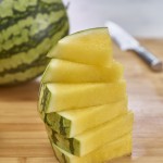 Six yellow watermelon triangular cuts with mini melon and knife in background set on bamboo cutting board