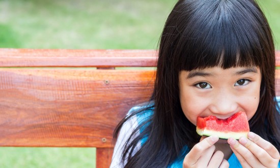 young Asian girl eating small watermelon slice, sitting on wooden bench, greenery in background