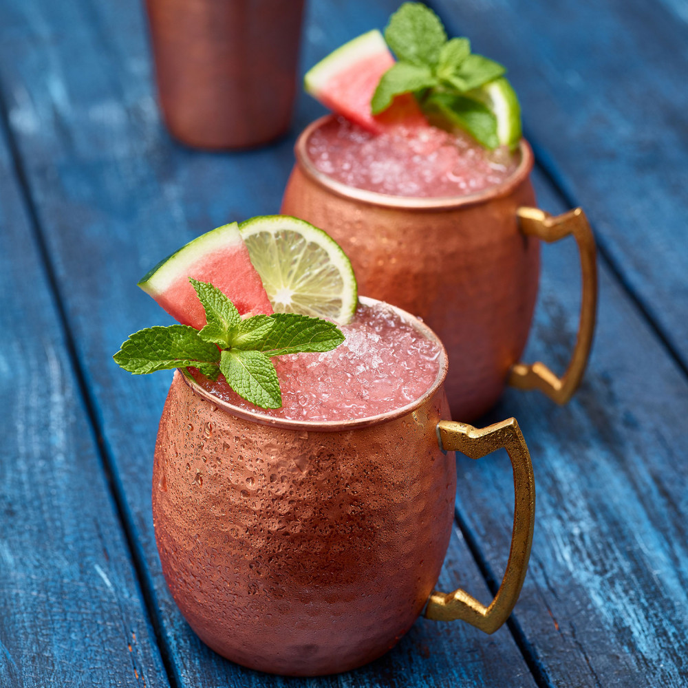 Watermelon Mule (x2) in copper cups garnished with watermelon slice, lime and mint. Copper shaker in background. Set on blue painted table