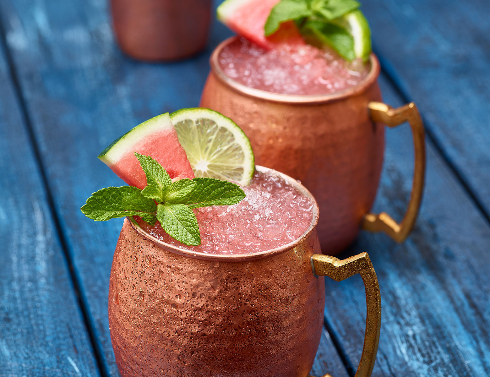 Watermelon Mule (x2) in copper cups garnished with watermelon slice, lime and mint. Copper shaker in background. Set on blue painted table