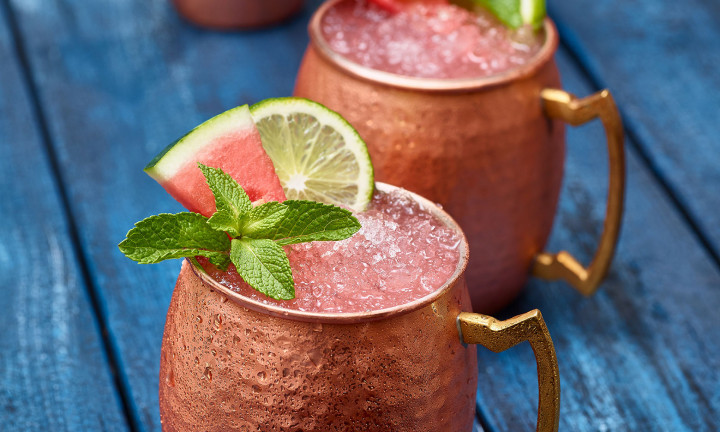 Watermelon Mule (x2) in copper cups garnished with watermelon slice, lime and mint. Copper shaker in background. Set on blue painted table