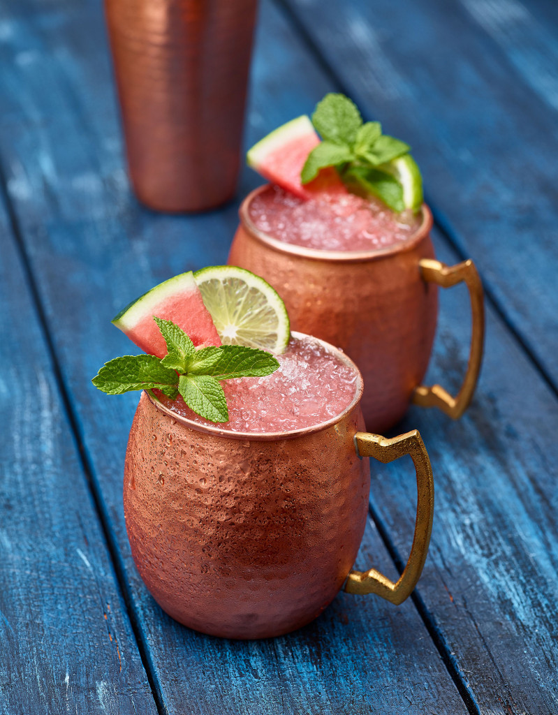 Watermelon Mule (x2) in copper cups garnished with watermelon slice, lime and mint. Copper shaker in background. Set on blue painted table