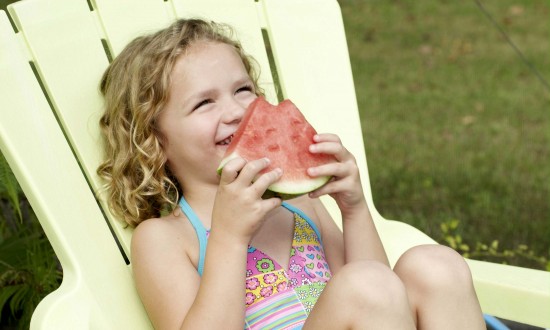 young girl in swimsuit in yellow adirondack chair smiling and eating watermelon slice