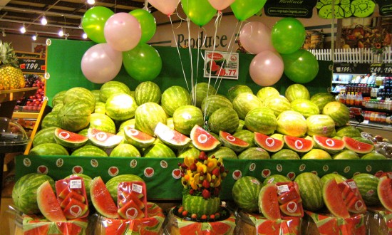 grocery store display contest set-up, whole watermelon, wedges, slices with watermelon carved basket in front/center. Pink and green balloons floating overhead.