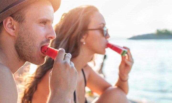 Man and woman, side-view, eating watermelon wedges (at beach)