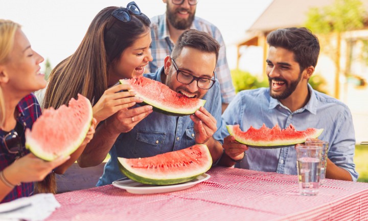 group of friends, three men, two women at outdoor picnic table wit red and white gingham tablecloth, eating watermelon wedges