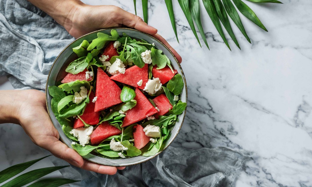 fresh summer watermelon salad held by two hand with palm leaves on sides. Set on gray and white marble