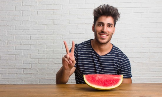 man at table with wedge of watermelon in front of him. He's making peace sign