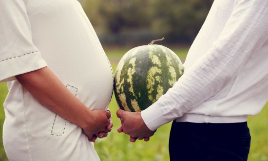 man and women standing facing each other. Women is pregnant. Man holding watermelon between them