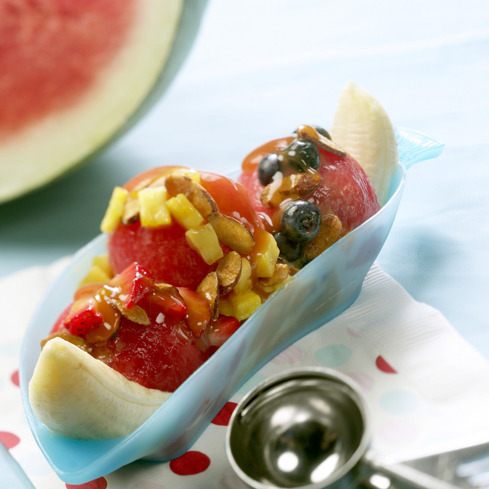 Watermelon Banana Split set in blue banana split dish. Dish set on white and red polka-dot paper napkin. Ice cream scoop in foreground. Watermelon wedge in background.