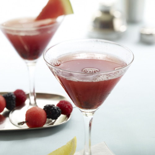 Two Watermelon Blackberry Martinis shown with skewered fruit and garnished with triangular watermelon cut and wedge of lemon alongside.