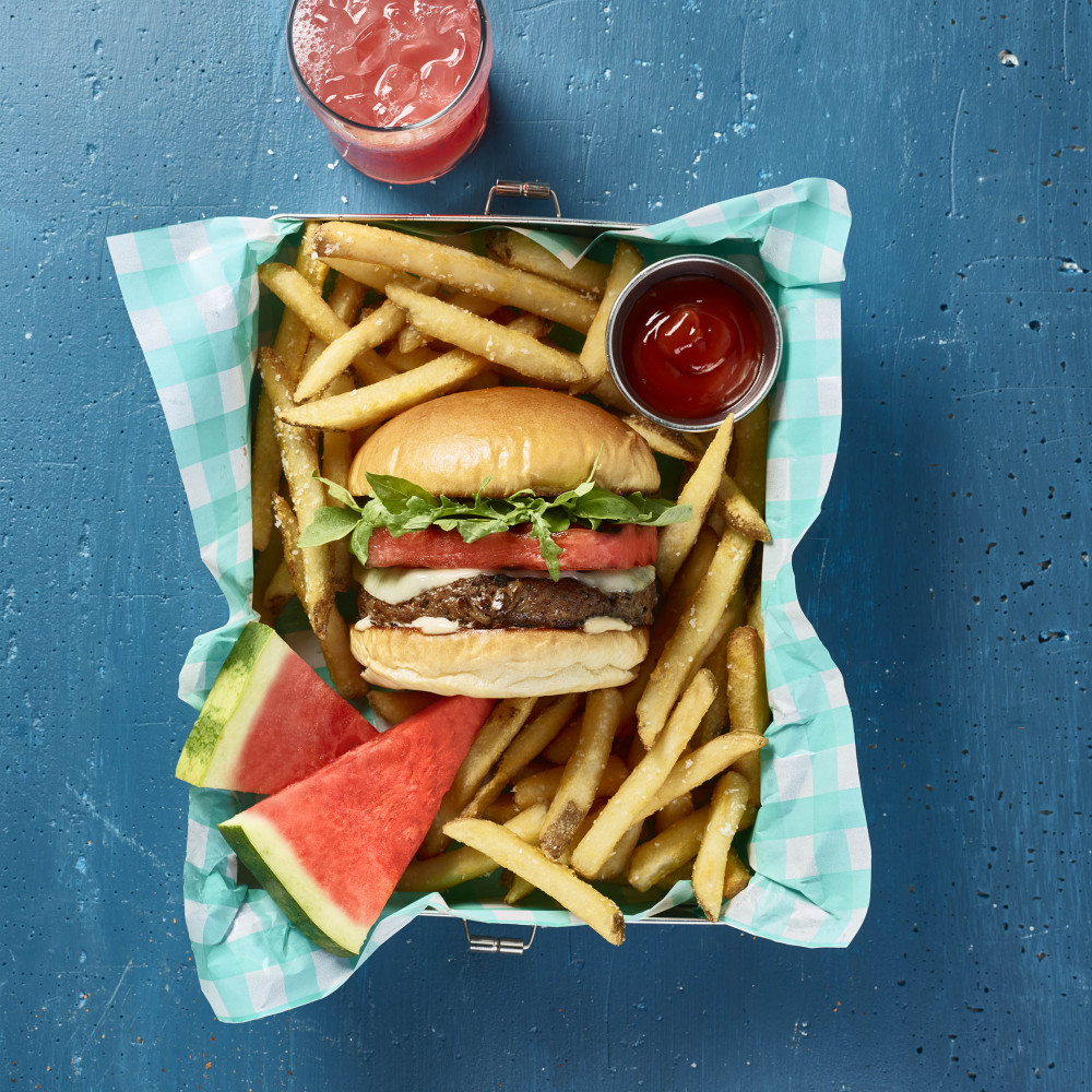 Hamburger served in paper lined serving basket. Hamburger is in center of french fries with side of ketchup. Two watermelon triangle pieces on side. Iced watermelon aqua fresca on side.