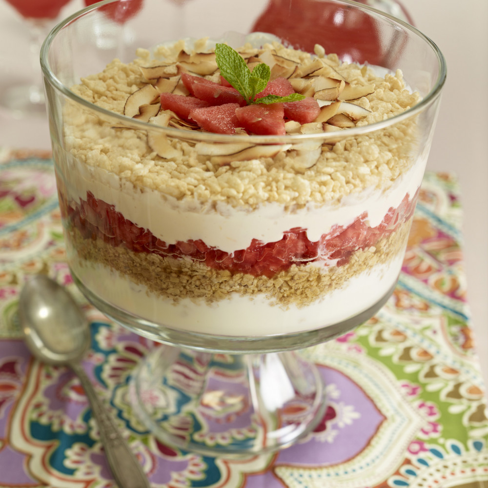 Watermelon Six Layer Trifle in trifle serving dish, garnished with shredded coconut, watermelon and mint leaves. Watermelon aqua fresca in pitcher and various types of glasses in background.