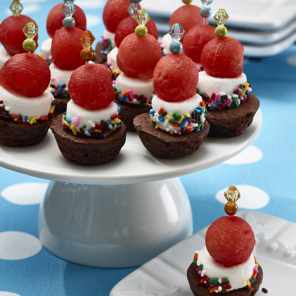 Dozen brownie bites set on white cake plate topped with decorative toothpicks. One brownie bite served on small dessert plate with more serving plates in background. Setting is aqua with white polka-dot tablecloth.